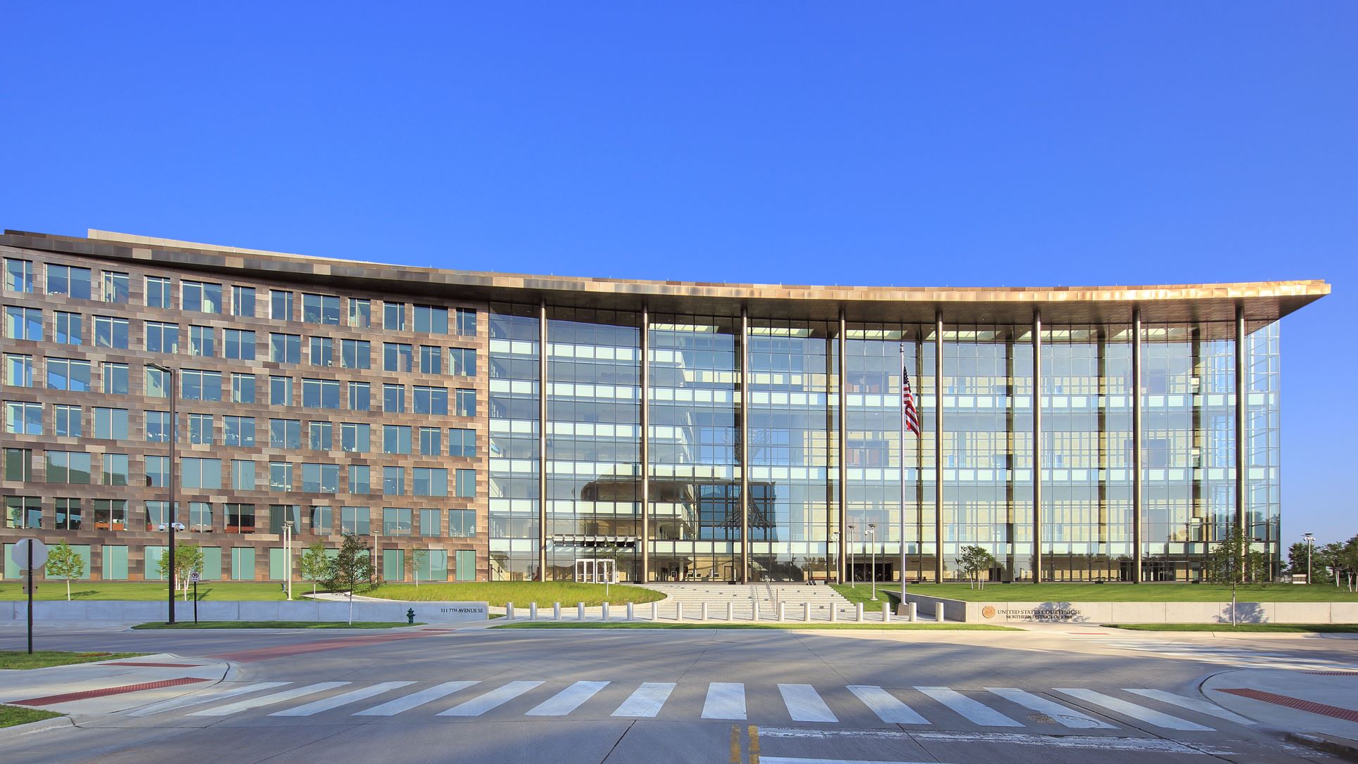 A multi-story beige building with glass windows across half of the front and a U.S. flag on a flagpole out front. Text on front wall: United States Courthouse