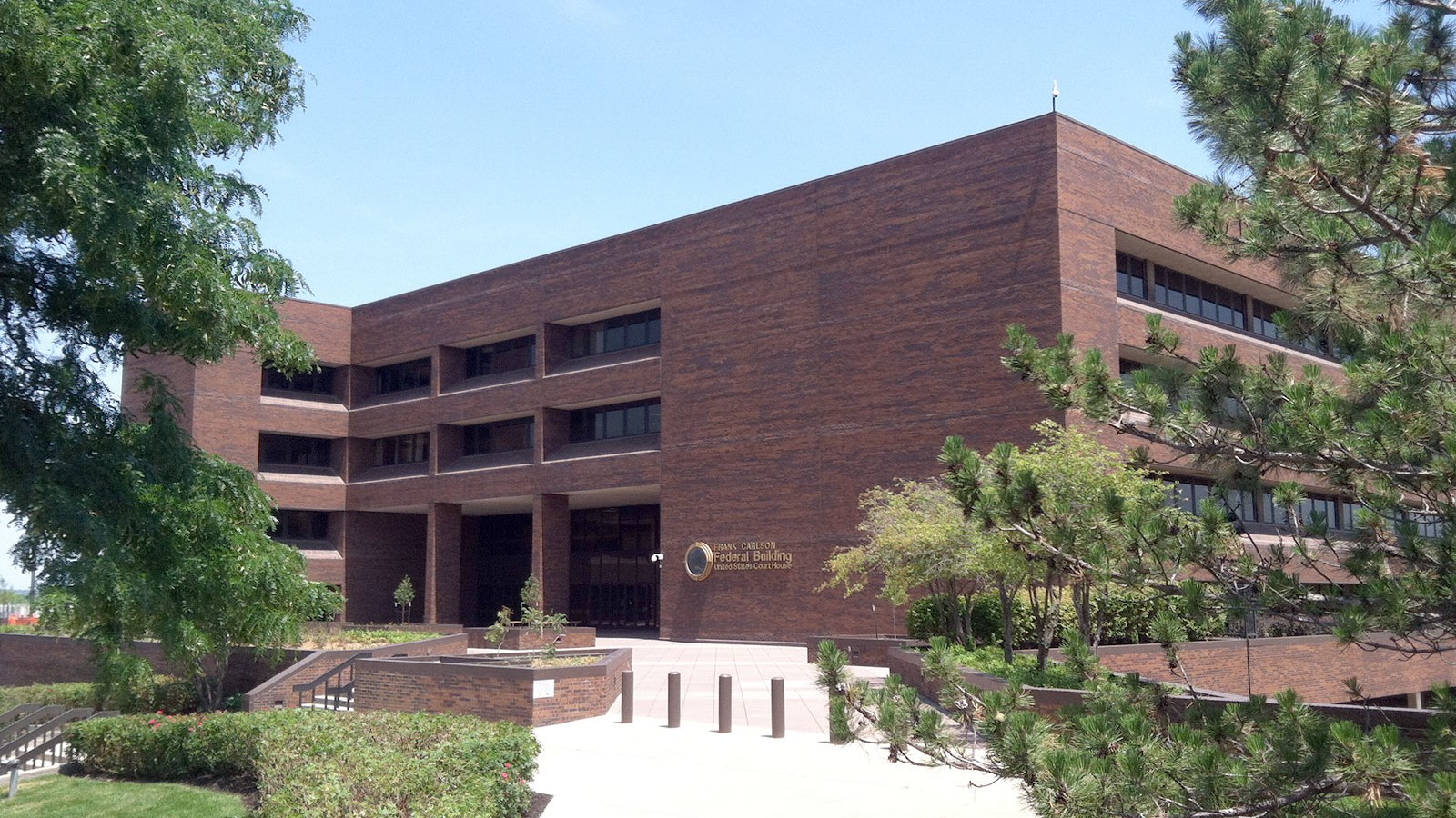 A red brick 3-level building framed by grass, trees and blue sky