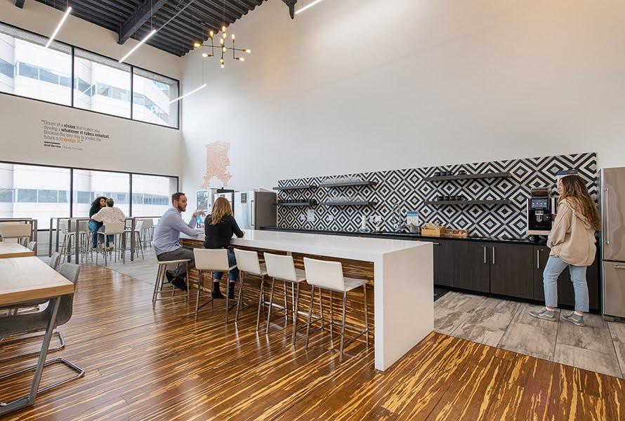 An office space with wood grained patterned floor a kitchen with several refrigerators and shelves with a diamond pattern backdrop and bar.